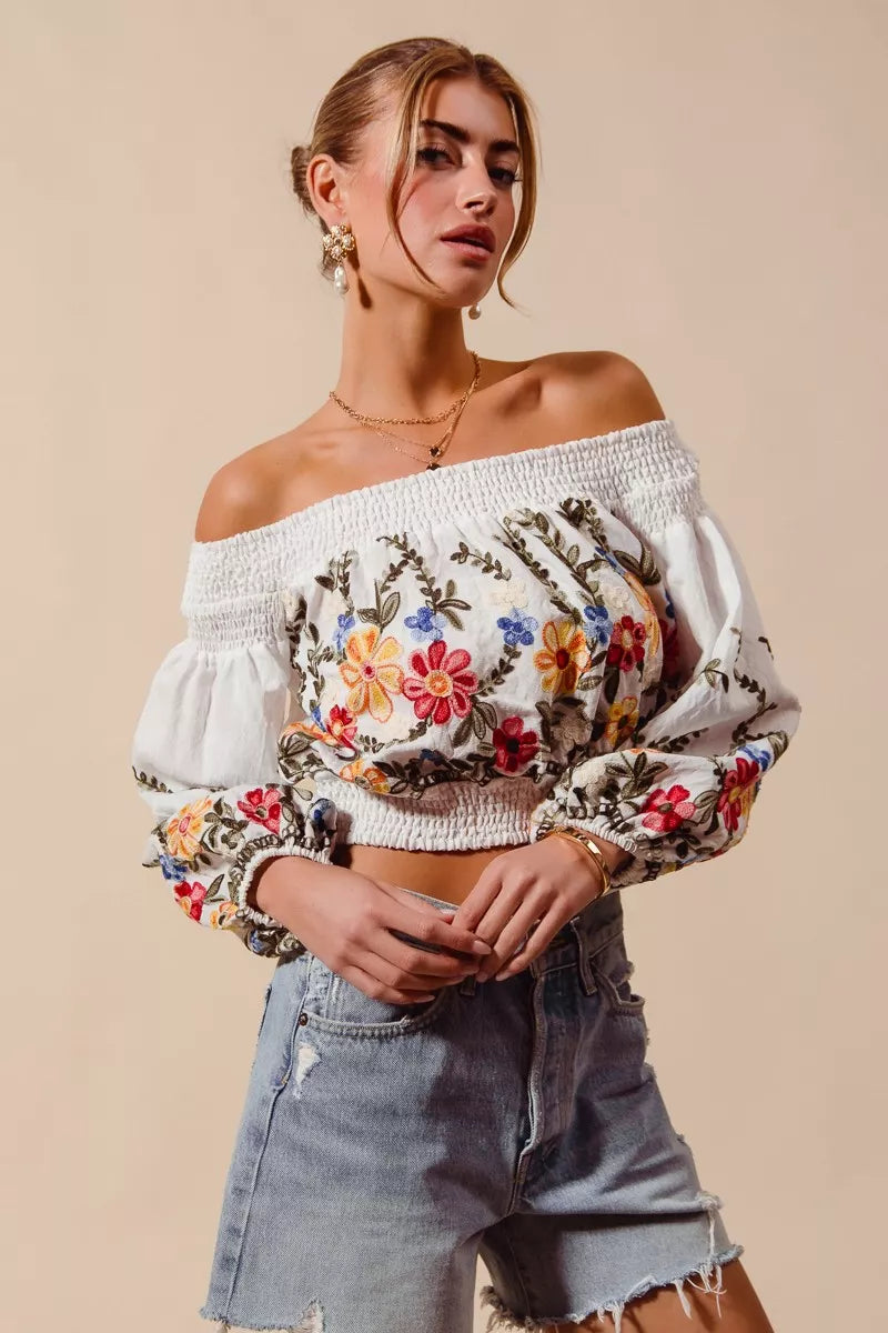 Woman wearing a floral off-shoulder top and denim shorts against a beige background