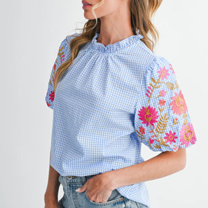 Woman wearing a blue checkered blouse with floral embroidery on a plain background