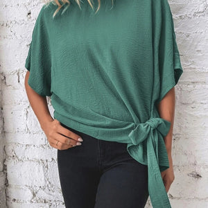 Person wearing a green top with a knot detail against a white brick wall.