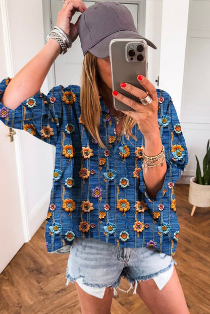 Person taking a mirror selfie wearing a blue floral blouse and denim shorts indoors.