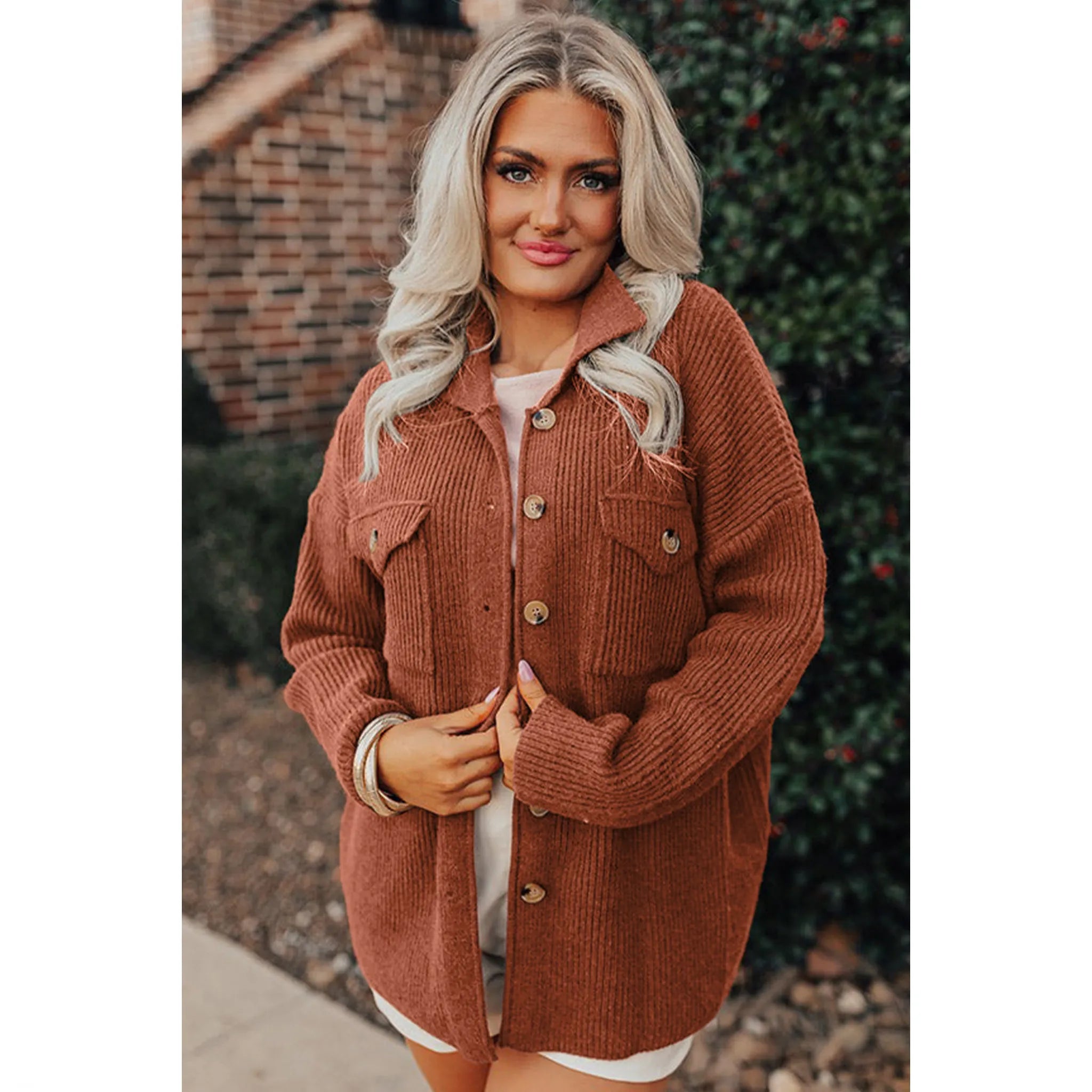 Woman wearing a brown corduroy jacket outdoors with a brick wall and greenery in the background.