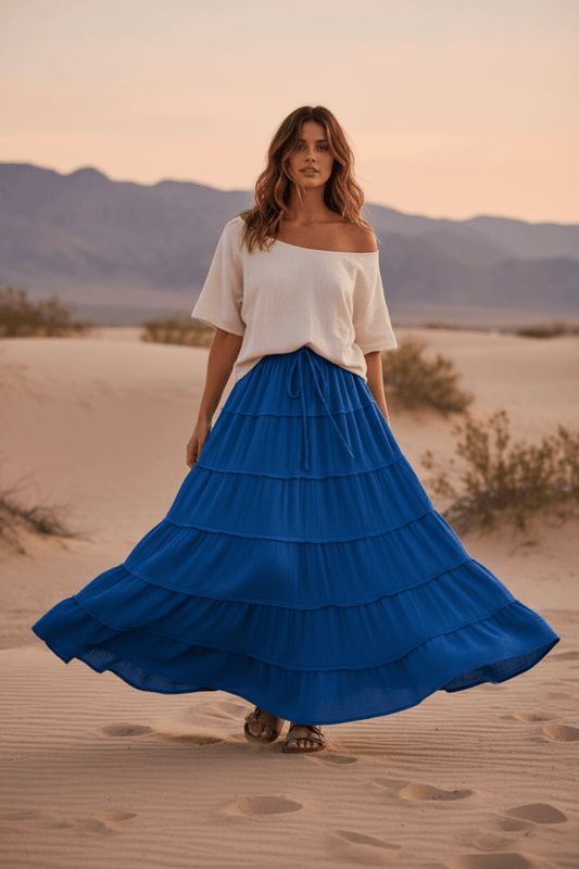 Woman in a blue skirt and white top standing in a desert landscape with mountains in the background.
