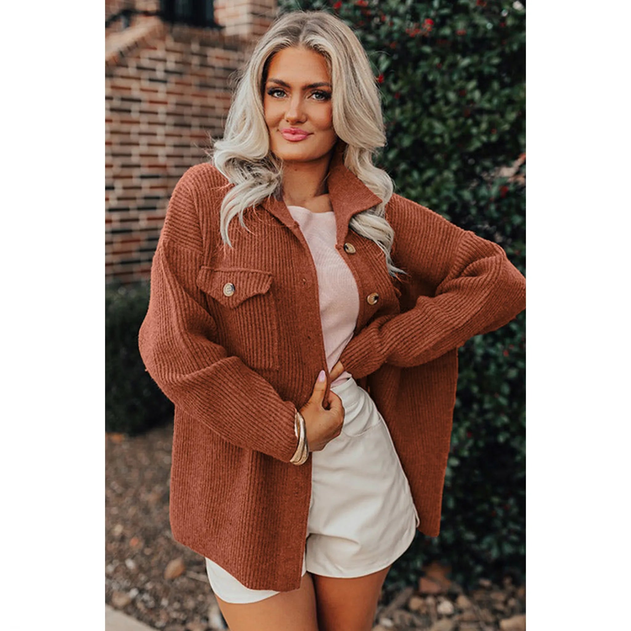 Woman wearing a brown cardigan over a white shirt and shorts, standing outdoors.