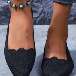 Black flat shoes with scalloped edges worn by a person on a textured surface.